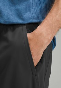 Sorte atletiske shorts med en sidelomme og glat stof. Toppen er en blå, struktureret skjorte. Hænderne er afslappet placeret i lommen.