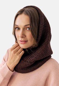Brązowy dzianinowy komin o grubym, teksturowanym wzorze. Noszony luźno, układa się wokół szyi i głowy na jasnym tle.