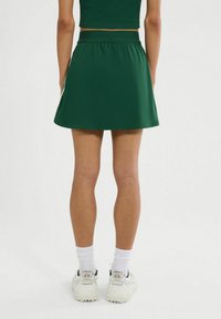 Zielona, rozszerzana spódnica wykonana z gładkiego materiału, z dopasowaną talią i prostym designem. Połączona z białymi skarpetkami i sportowymi butami.