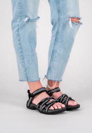 Schwarze und graue gewobene Sandalen mit strukturiertem Obermaterial und verstellbaren Riemen, kombiniert mit ausgebeulten hellblauen Jeans mit fransigen Säumen.