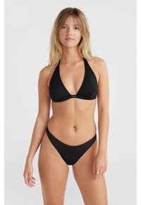 Young woman with blonde hair wearing a black halter bikini top and matching bikini bottoms standing against a plain light background.