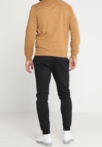 Felpa marrone con collo e polsini a costine, abbinata a joggers neri a gamba stretta. I joggers hanno una superficie liscia e una vita elasticizzata.