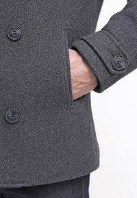 Szary wełniany płaszcz z guzikiem i boczną kieszenią. Materiał wydaje się mieć fakturę, a guziki są ozdobione nazwą marki.