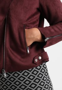 Giacca bordeaux in finta pelle scamosciata con cerniere argentate, tasche frontali inclinate e dettaglio polsini con bottoni, abbinata a una gonna nera e bianca con motivo a fantasia.