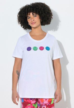 Kobieta z kręconymi włosami w białej koszulce z czterema kolorowymi buźkami i żywymi, kwiatowymi wzorzystymi spodniach.