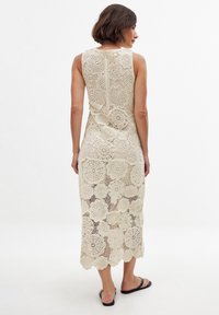 Knee-length, sleeveless dress in beige crocheted lace with floral patterns, scalloped hem, and a straight back design. Flat black sandals worn.