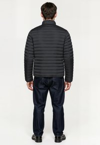 Black puffer jacket with horizontal quilted design, high collar, worn over dark denim pants. Black shoes complete the outfit.