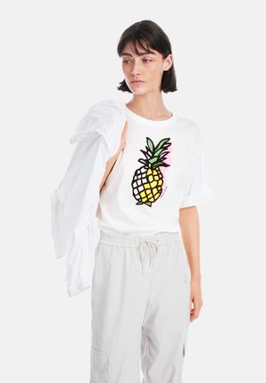 Weißes T-Shirt mit kurzen Ärmeln, das ein buntes Ananas-Graphic zeigt, kombiniert mit hellgrauen elastischen Hosen und einer weißen Jacke, die über die Schulter drapiert ist.
