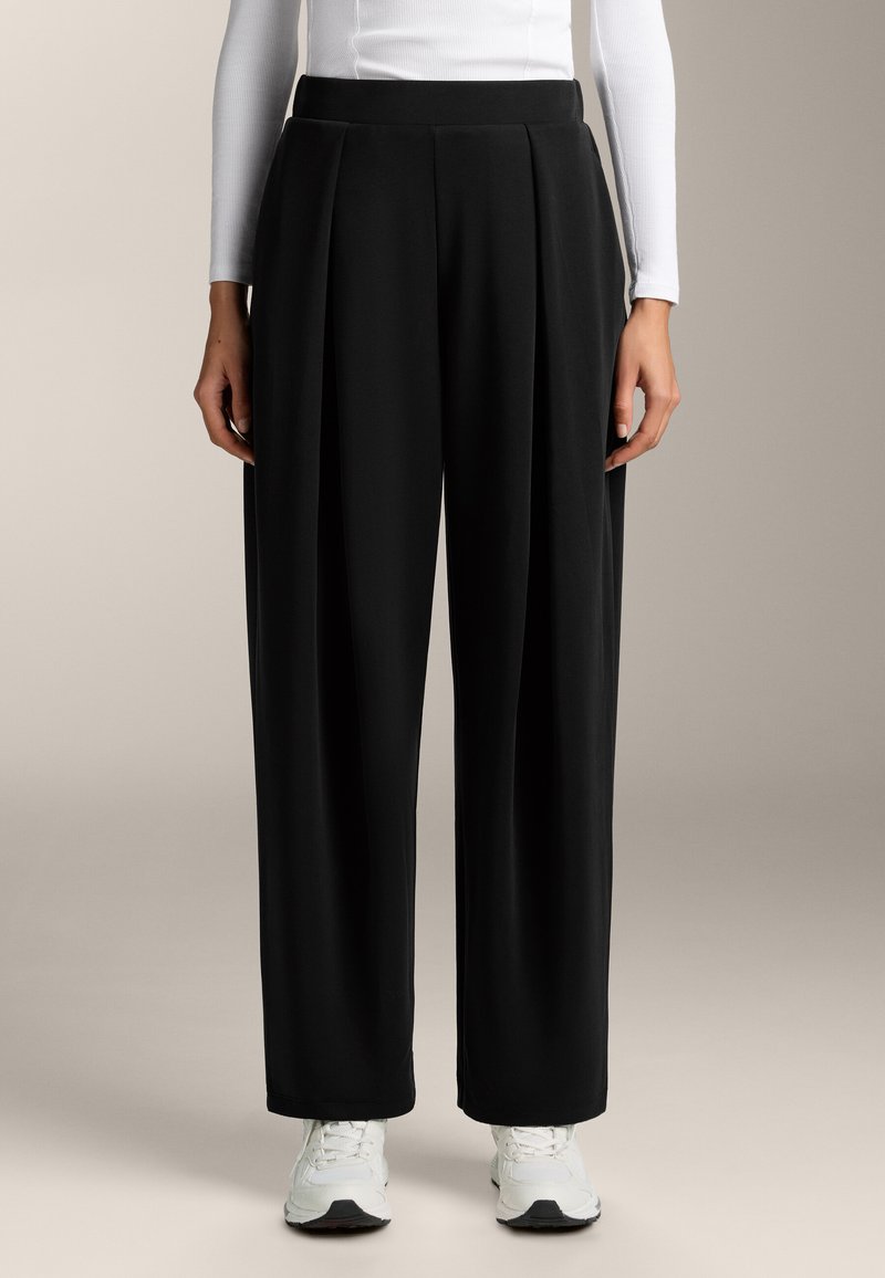 Fekete, rakott, bő lábú nadrág, puha anyagból készült, magas derékkal, sima felülettel és laza fazonnal. Fehér sportcipővel kiemelve.