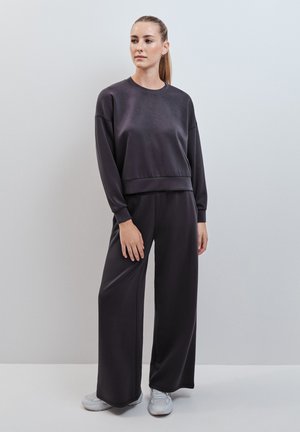Donna con capelli raccolti che indossa una felpa larga e scura e pantaloni a gamba larga con sneakers bianche, in piedi contro un muro grigio chiaro.