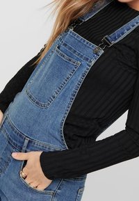 Lichte blauwe denim salopette met verstelbare schouderbanden, een voorzak en zijzakken, gecombineerd met een zwart ribbelshirt met lange mouwen.