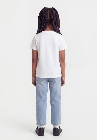 Παιδί με μακριά πλεξούδα που φοράει απλό λευκό T-shirt, ανοιχτόχρωμο μπλε τζιν, λευκές κάλτσες και μαύρα αθλητικά παπούτσια, στέκεται μπροστά σε λευκό φόντο.
