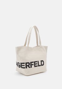Beżowa torba z płótna z dwiema rączkami i dużym czarnym napisem częściowo widocznym "AGERFELD" z przodu.