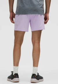 Shorts de sport violet clair en matériau léger, avec une texture douce. Portés avec des baskets noires et des chaussettes blanches.