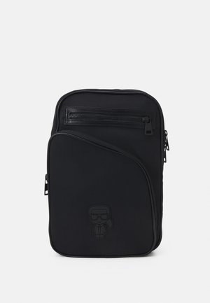 Sac à dos noir en matériau lisse, avec une poche zippée à l'avant, un design incurvé et un détail de caractère en relief sur le bas à l'avant.