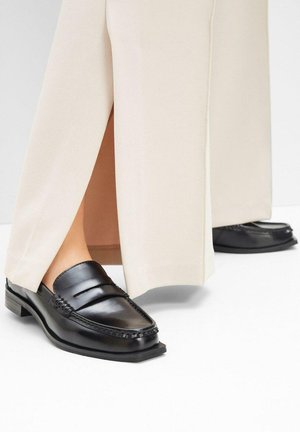 Mocassini in pelle nera con punta arrotondata e cuciture decorative, abbinati a pantaloni beige a gamba larga che coprono parzialmente le scarpe.