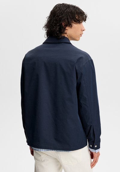 Uomo con capelli ricci che indossa una giacca blu scuro sopra una camicia a righe e pantaloni bianchi, visto di spalle e che guarda di lato.