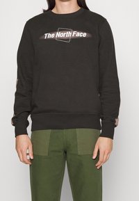 Svart sweatshirt med rund hals, med vit "The North Face"-logotyp. Långa ärmar med ljusbruna detaljer. Matchas med gröna byxor.