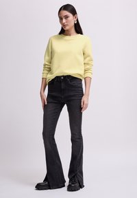 Maglione giallo struttura a trama con scollatura rotonda abbinato a jeans neri a vita alta con fianchi svasati e aperture laterali, indossato con stivali neri.