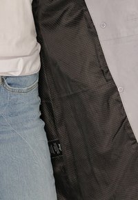 Lichtgrijze jas met knopen. Interieur heeft een donkere stof met kleine witte stippen. Draagt over casual denim.