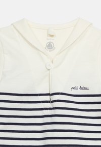 Λευκό βρεφικό πουκάμισο με ναυτικό μπλε οριζόντιες ρίγες, ένα λευκό κουμπί κοντά στον γιακά και το «petit bateau» κεντημένο στο στήθος.
