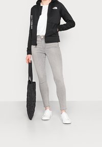 Sudadera con cremallera negra y capucha, jeans ajustados grises, zapatillas blancas y una bolsa tote negra acolchada, con una textura de tela suave.