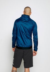 Man som vänder ryggen mot kameran, klädd i en blå huvjacka av vindtåligt material och svarta shorts med vit text, mot en enkel vit bakgrund.