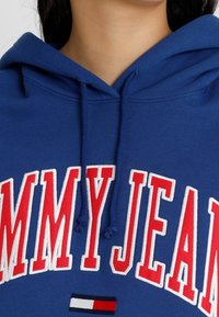 Sudadera de algodón azul con gran texto en blanco y rojo "TOMMY JEAN" en el frente. Incluye capucha con cordón y acento de logo en contraste.