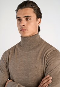 Brun polotröja i texturerat stickat tyg, med hög hals och subtil logodetalj på bröstet.