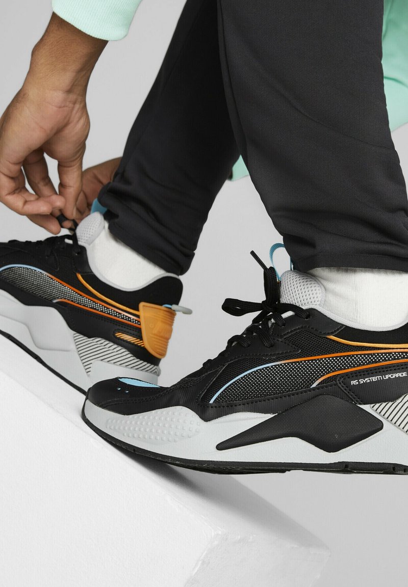 Personne portant des baskets noires, blanches et orange en train de lacer ses chaussures, debout sur une surface blanche inclinée, avec un pantalon noir et des chaussettes blanches.