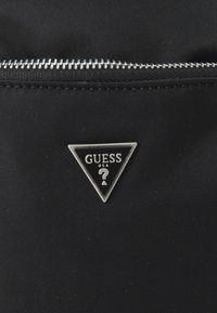 Tela de nylon negra con un zipper plateado. Presenta una placa triangular con el logo de Guess, que incluye el nombre de la marca y un signo de interrogación en el centro.
