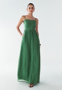 Zaļa maksi kleita ar teksturētu, krokotu dizainu, kurai ir plānas siksnas un dekoratīvs viļņots apdare jostā un apakšmalā.