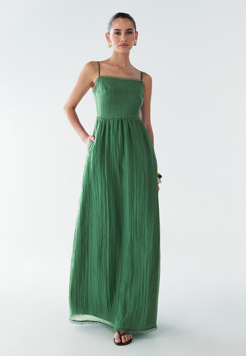 Zaļa maksi kleita ar teksturētu, krokotu dizainu, kurai ir plānas siksnas un dekoratīvs viļņots apdare jostā un apakšmalā.