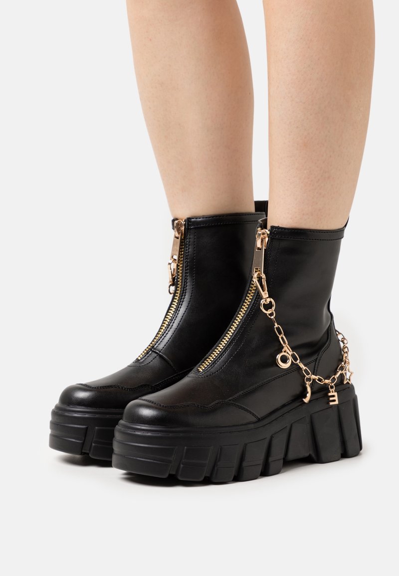 Bottines noires à plateforme épaisse avec fermetures éclair dorées à l'avant et chaîne décorative dorée avec lettres et charms étoiles.