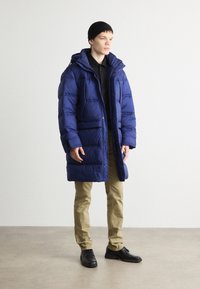 Cappotto imbottito blu con cappuccio, caratterizzato da sezioni trapuntate e tasche frontali, abbinato a pantaloni beige e scarpe nere, indossato da un modello.