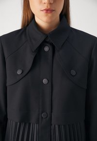 Femme portant un manteau noir boutonné avec des détails décoratifs à rabat sur les épaules et un tissu plissé sous la taille.