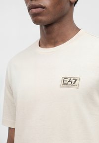Άνδρας που φοράει ένα απλό ανοιχτό μπεζ T-shirt με ένα μικρό λογότυπο EA7 Emporio Armani στο αριστερό στήθος.