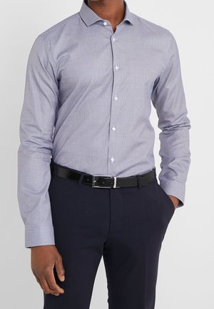 Mann trägt ein helllila gemustertes Button-up-Hemd, das in dunkle marineblaue Hosen mit einem schwarzen Ledergürtel gesteckt ist, eine Hand in der Tasche.