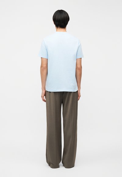 Άτομο με κοντά μαύρα μαλλιά που φοράει ανοιχτό μπλε κοντομάνικο πουκάμισο και φαρδιά καφέ παντελόνια, στέκεται με την πλάτη προς τον θεατή σε λευκό φόντο.