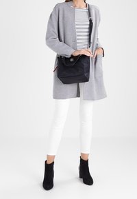 Kvinde iført grå frakke, stribet skjorte, hvide bukser, sorte ankelstøvler og holder en sort skuldertaske med et logo.