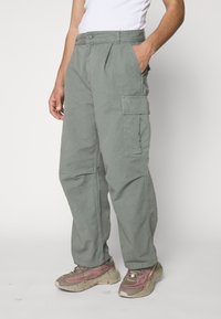 Spodnie typu cargo w stonowanym oliwkowym kolorze, wykonane z bawełny. Posiadają boczne kieszenie, luźny krój i minimalne przeszycia. Połączone z masywnymi, neutralnymi sneakersami.