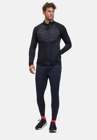 Sportliche Herrenjacke in Schwarz mit gesteppter Oberpartie; tailliertes Design, glatter Stoff, Reißverschluss, kombiniert mit dunklen Leggings und roten Akzentsocken.
