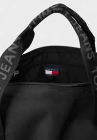 Czarna torba z materiału z zamkiem błyskawicznym na górze, z metką z logo wewnątrz oraz czarnymi uchwytami z tekstem "TOMMY JEANS" wtopionym w materiał.