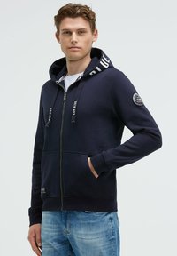 Sudadera con cremallera color navy, con cordones, detalles a rayas en la capucha y bolsillos frontales; incluye un parche con el logo en la manga.