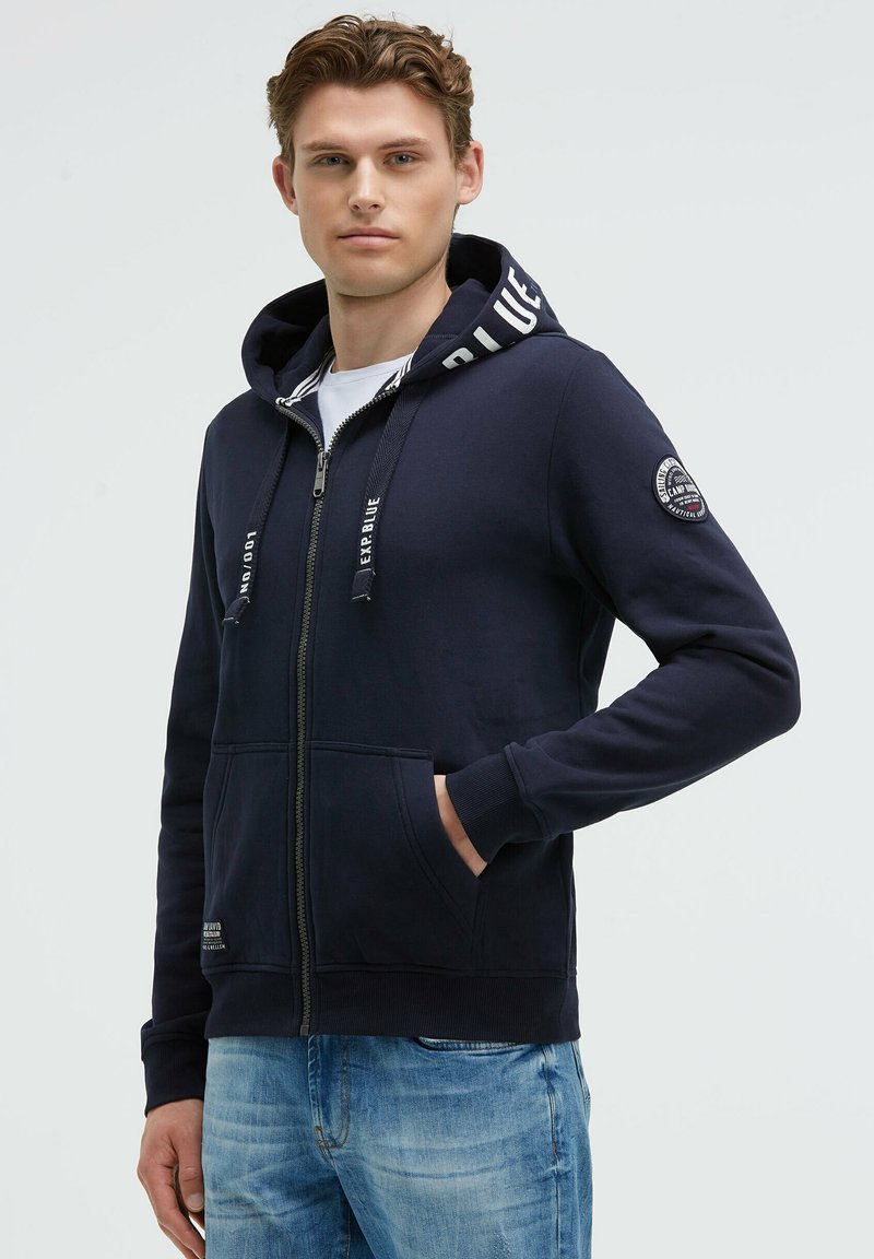 Sudadera con cremallera color navy, con cordones, detalles a rayas en la capucha y bolsillos frontales; incluye un parche con el logo en la manga.