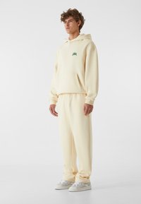 Cremefarvet hoodie og matchende sweatpants, blødt materiale, ribbede manchetter, frontlomme, grøn logo-detalje på brystet, parret med lyse sneakers.