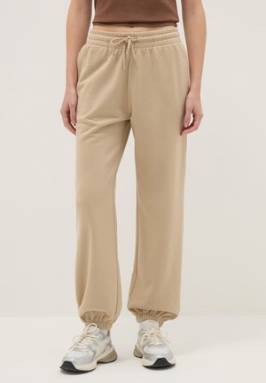 Pantalones jogger beige con cintura elástica, cordón y puños elásticos, usados con zapatillas blancas y plateadas por una persona de pie.