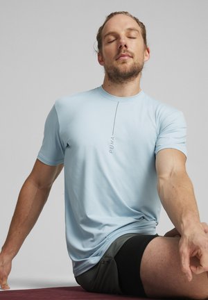 Světle modré krátké sportovní tričko s minimalistickým designem, které má vertikální černé logo. Model sedí v jogové pozici na podložce.