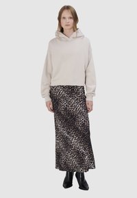 Vrouw draagt een beige hoodie, een lange rok met luipaardprint en zwarte enkellaarzen, staand tegen een effen lichte achtergrond.