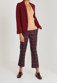 Blazer bordeaux, top enroulé beige, et pantalon à carreaux marron avec des lignes bleues. Chaussures bordeaux brillantes. Coupe ajustée et texture de tissu lisse.